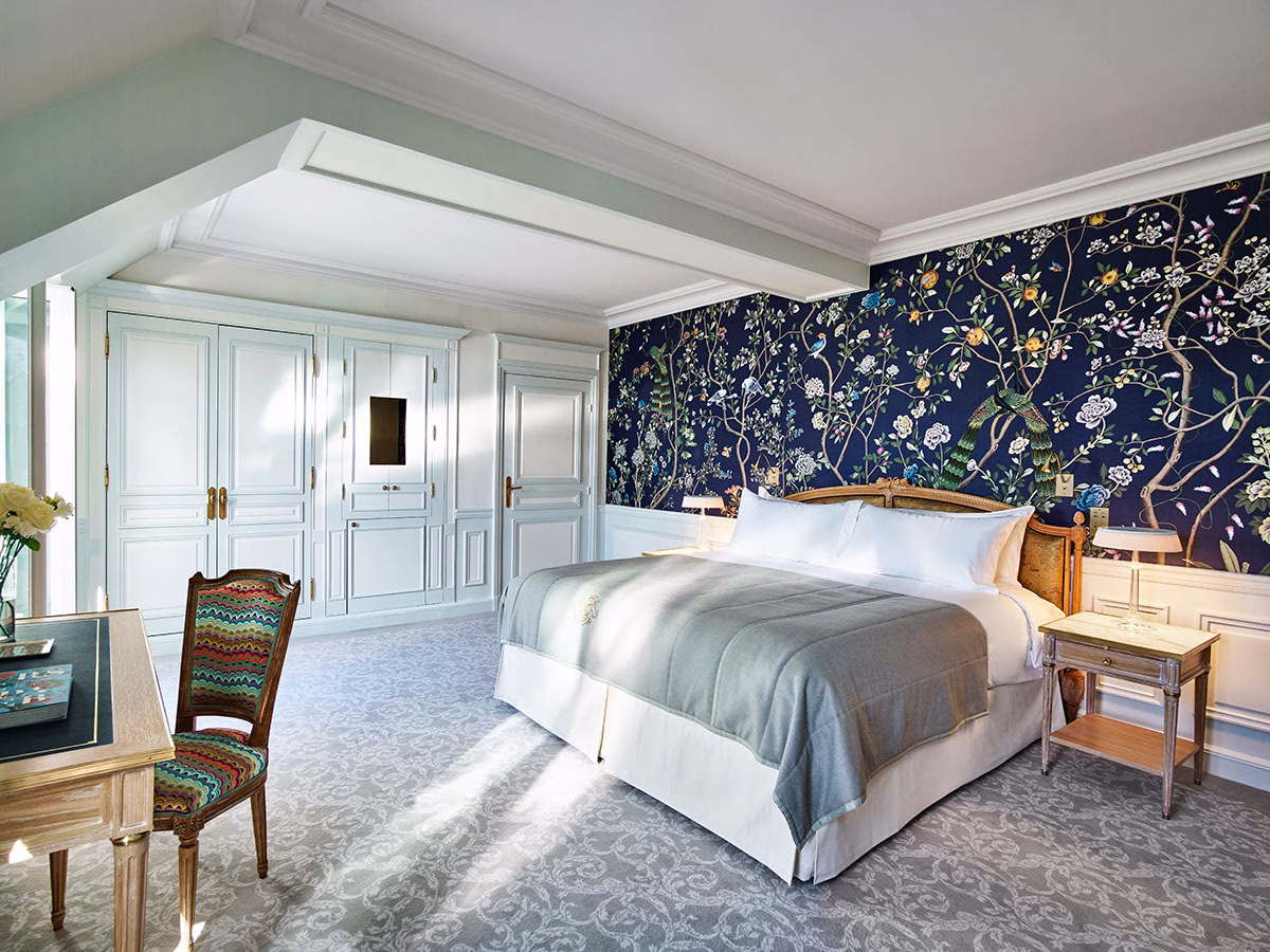Ein herrschaftliches Schlafzimmer mit einem großen Kingsize-Bett vor einer tiefblauen, kunstvollen Tapete mit floralen Mustern und Vögeln, kombiniert mit weißen Holzschränken und einem verzierten Schreibtischstuhl im Retro-Look