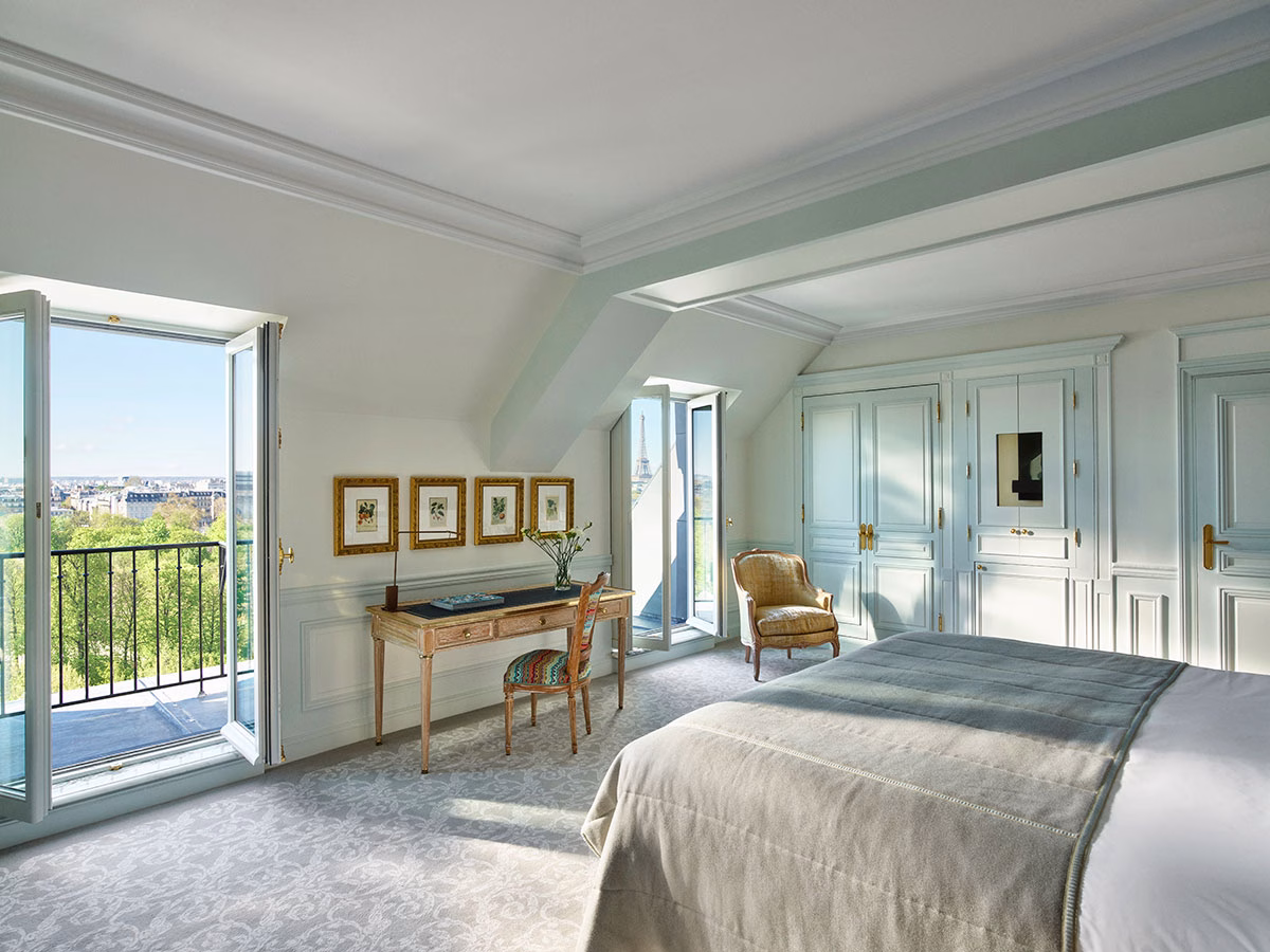 Das lichtdurchflutete Schlafzimmer einer Dachsuite im Le Meurice mit geöffneten Balkontüren, die den Blick auf den Pariser Eiffelturm freigeben, ausgestattet mit einem eleganten Holzschreibtisch und hellblauen Einbauschränken