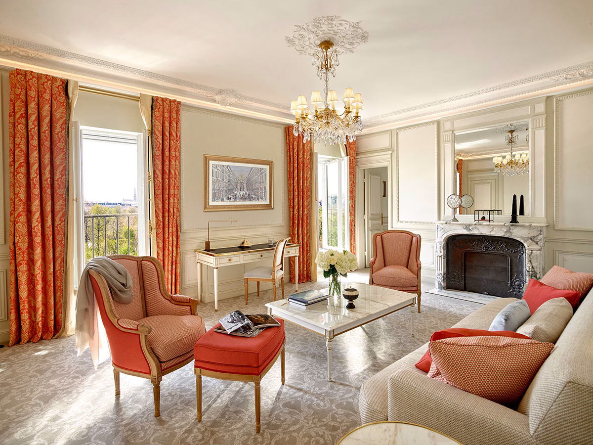 Ein prachtvolles Wohnzimmer im klassischen Pariser Stil mit einem weißen Marmorkamin, einem Kristallleuchter, eleganten Ohrensesseln in Lachsrot, einem passenden Hocker und schweren, rot-gold gemusterten Vorhängen an den Fenstern