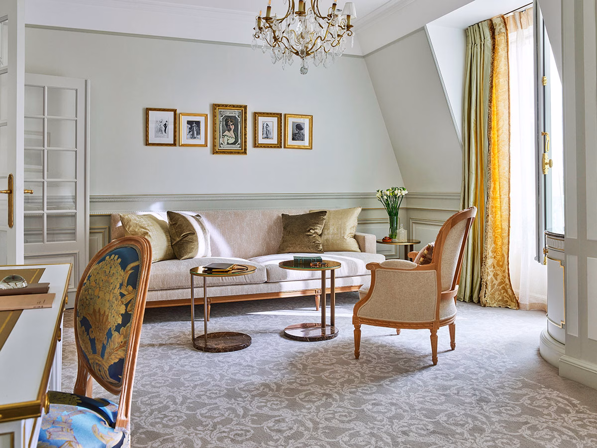 Eine stilvolle Suite-Sitzecke mit einem hellen Sofa, Samtkissen, runden Marmor-Beistelltischen und einem antiken Holz-Sessel auf einem grau gemusterten Teppich unter einem filigranen Kristallkronleuchter