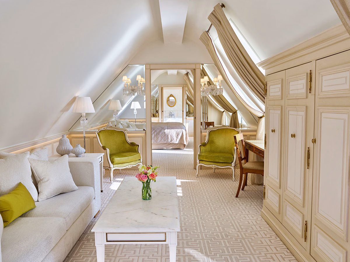 Das gemütliche Wohnzimmer einer Mansarden-Suite mit Dachschrägen, einem hellen Sofa, zwei olivgrünen Samtsesseln vor einer verspiegelten Wand und einem weißen Marmor-Couchtisch mit frischen Blumen