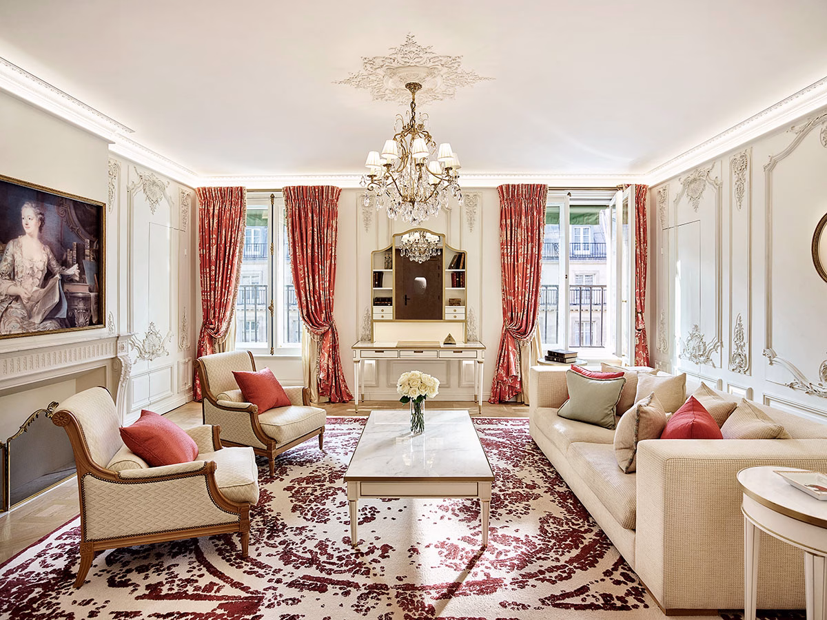 Ein lichtdurchfluteter, eleganter Hotelsalon im Barockstil mit verzierten Wandpaneelen, einem historischen Frauenporträt über dem Kamin, lachsfarbenen Sesseln, einem großen Sofa und edlen roten Vorhängen an den Fenstern