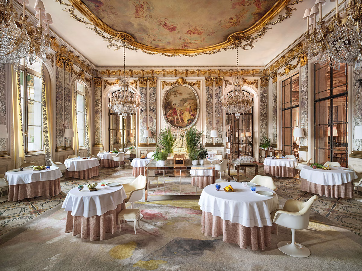 Der majestätische, von Versailles inspirierte Hotelsaal des Restaurant Le Meurice Alain Ducasse mit opulenten Kristallkronleuchtern, goldenen Deckenfresken, Marmorsäulen und fein eingedeckten, runden Tischen auf einem bunten Teppich