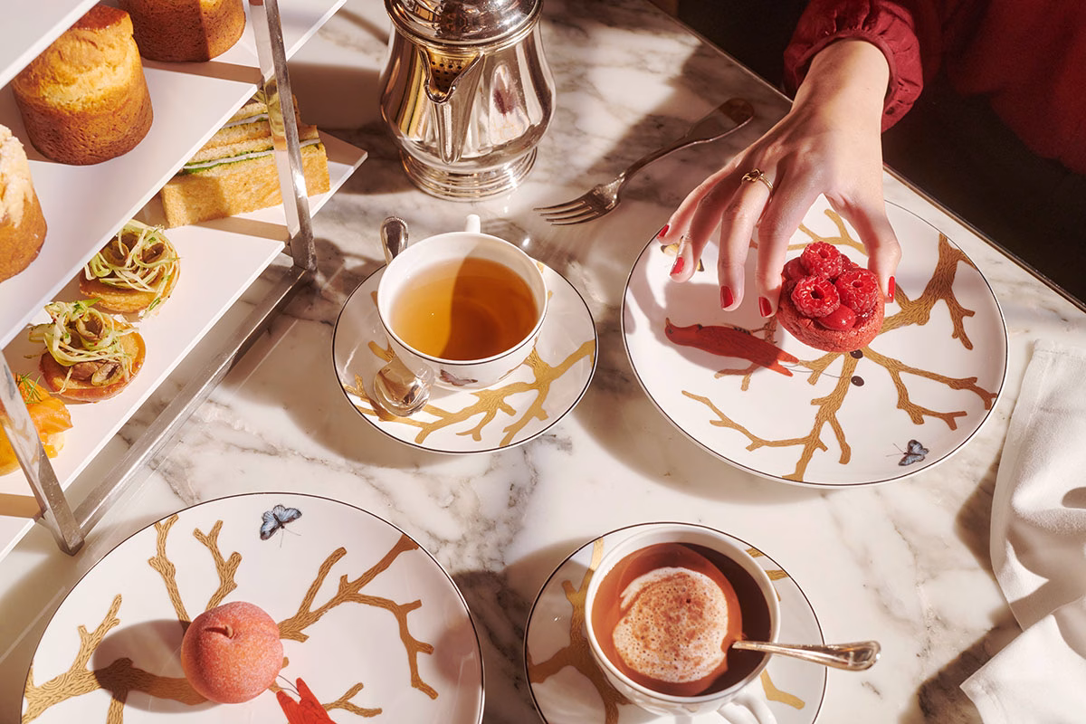 Draufsicht auf einen Marmortisch beim Afternoon Tea, auf dem fein verziertes Porzellan mit Ast-Motiven, eine Tasse Tee, heißer Kakao und eine Hand, die nach einem frischen Himbeertörtchen greift, angerichtet sind