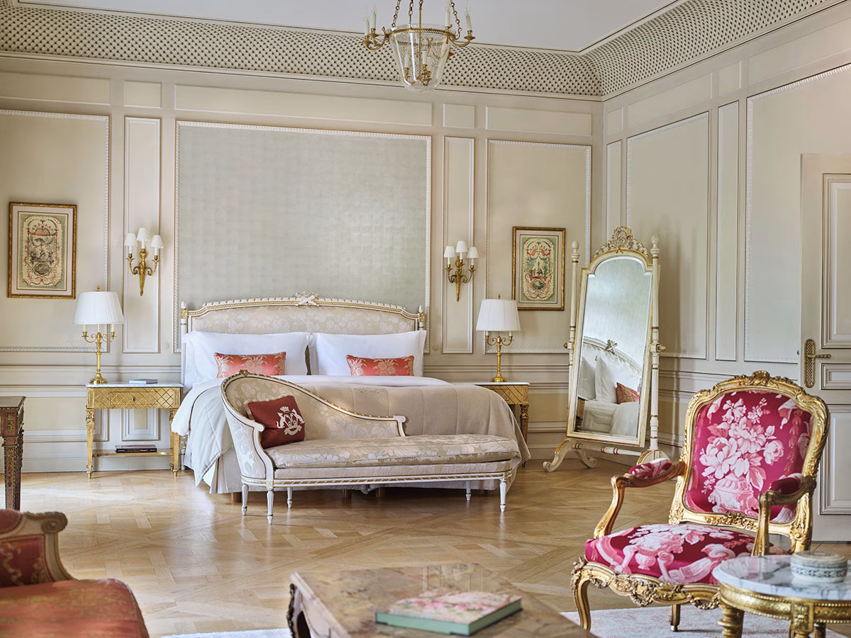 Ein luxuriöses, weitläufiges Hotelzimmer mit hellem Parkettboden, einem Kingsize-Bett mit zartgrünem Kopfteil, einer Chaiselongue am Fußende, einem großen barocken Standspiegel und einem auffälligen, pink-floral gemusterten Sessel im Vordergrund
